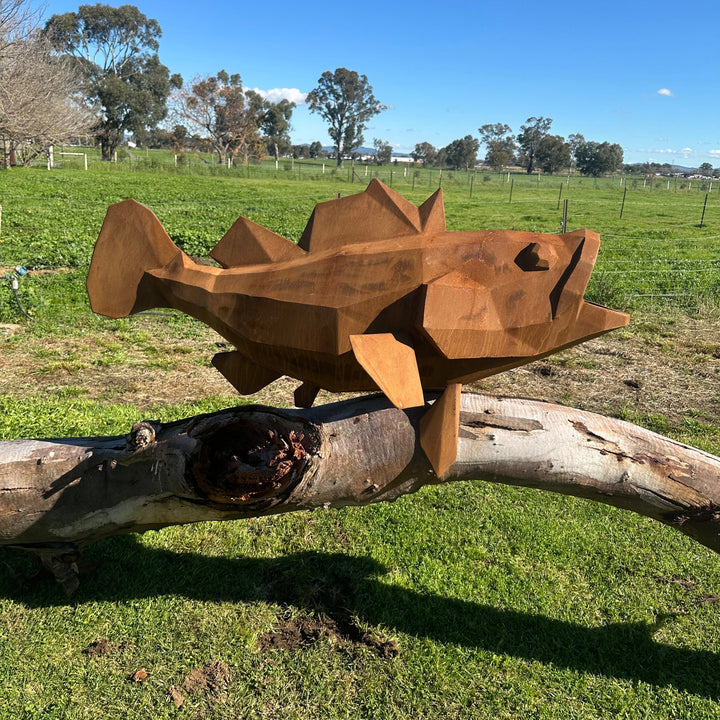 A geometric, faceted metal sculpture of a fish resting on a weathered log, set outdoors in a grassy field under a clear blue sky.